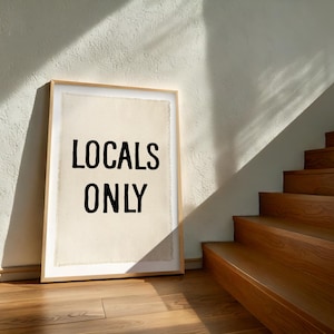 Pode incluir: Uma impress&atilde;o emoldurada com o texto "LOCALS ONLY" em letras pretas. A impress&atilde;o est&aacute; encostada a uma parede branca, com uma escada de madeira ao fundo. A impress&atilde;o tem um fundo claro e uma moldura de madeira.