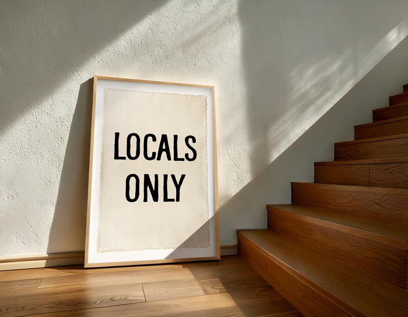 Pode incluir: Uma impress&atilde;o emoldurada com o texto "LOCALS ONLY" em letras pretas. A impress&atilde;o est&aacute; encostada a uma parede branca, com uma escada de madeira ao fundo. A impress&atilde;o tem um fundo claro e uma moldura de madeira.