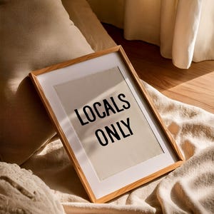 Pode incluir: Uma impress&atilde;o emoldurada com o texto "LOCALS ONLY" em letras pretas. A moldura &eacute; feita de madeira clara, e a impress&atilde;o est&aacute; em um fundo branco. A impress&atilde;o est&aacute; em cima de um cobertor bege.