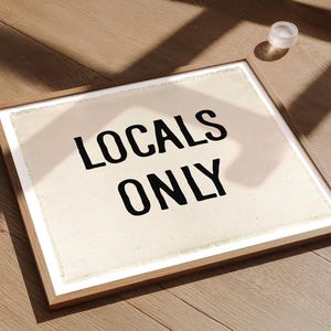 Pode incluir: Uma obra de arte emoldurada com o texto "LOCALS ONLY" em letras pretas em negrito sobre um fundo creme. A moldura &eacute; de cor de madeira clara. Um pequeno objeto redondo branco est&aacute; perto da moldura em uma superf&iacute;cie de madeira.