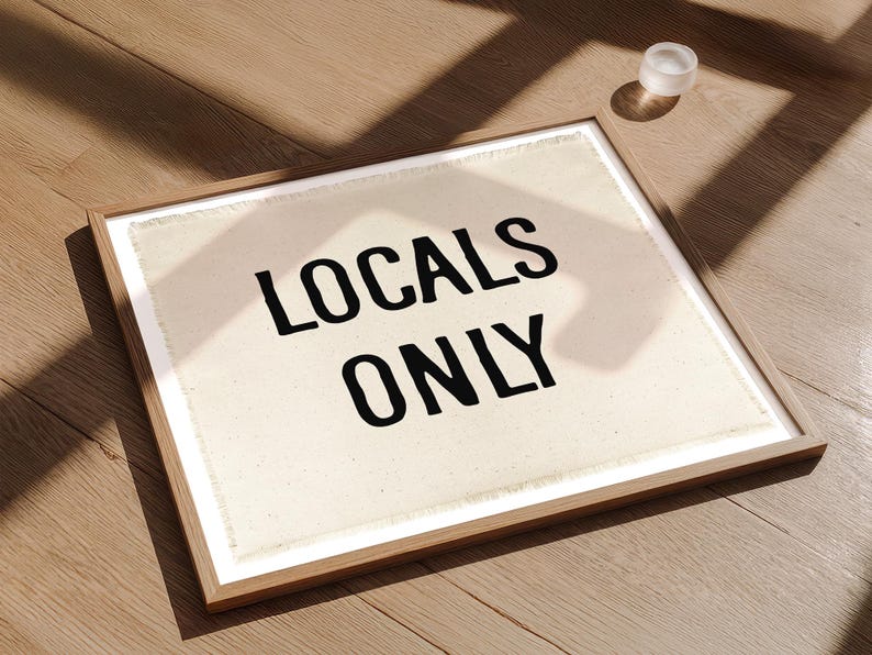 Pode incluir: Uma obra de arte emoldurada com o texto "LOCALS ONLY" em letras pretas em negrito sobre um fundo creme. A moldura &eacute; de cor de madeira clara. Um pequeno objeto redondo branco est&aacute; perto da moldura em uma superf&iacute;cie de madeira.