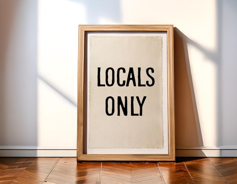 Pode incluir: Uma impress&atilde;o emoldurada com as palavras "LOCALS ONLY" em letras pretas em negrito. A impress&atilde;o tem um fundo texturizado esbranqui&ccedil;ado e est&aacute; emoldurada em uma moldura de madeira marrom clara. A obra de arte est&aacute; encostada em uma parede branca em um ch&atilde;o de madeira.