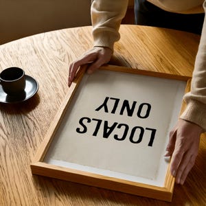 Pode incluir: Uma tela emoldurada com o texto "LOCALS ONLY" em preto, exibida em uma mesa de madeira. A moldura &eacute; feita de madeira clara. Uma x&iacute;cara e um pires de caf&eacute; pretos est&atilde;o na mesa.