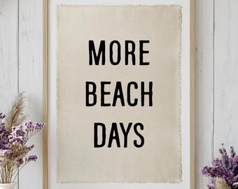 May include: A framed wall art print featuring a light beige fabric background with frayed edges. The text MORE BEACH DAYS is printed in bold black capital letters centered on the fabric. The piece is enclosed in a simple light wood frame.