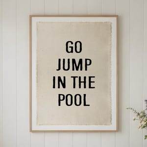 Peut inclure: Une décoration murale rectangulaire composée d'une toile en tissu beige clair aux bords effilochés, encadrée dans un cadre en bois clair. La toile présente le texte noir GO JUMP IN THE POOL en lettres majuscules grasses et sans empattement.