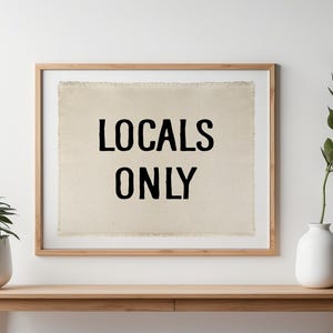 Pode incluir: Uma impress&atilde;o emoldurada com o texto "LOCALS ONLY" em preto sobre um fundo bege. A moldura &eacute; marrom claro. Uma pequena planta em vaso e um vaso com vegeta&ccedil;&atilde;o est&atilde;o em uma mesa de madeira.