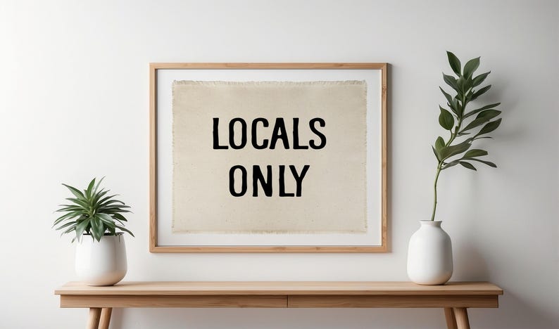 Pode incluir: Uma impress&atilde;o emoldurada com o texto "LOCALS ONLY" em preto sobre um fundo bege. A moldura &eacute; marrom claro. Uma pequena planta em vaso e um vaso com vegeta&ccedil;&atilde;o est&atilde;o em uma mesa de madeira.