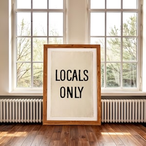 Pode incluir: Uma impress&atilde;o emoldurada em madeira com as palavras "LOCALS ONLY" em preto sobre um fundo creme. A impress&atilde;o est&aacute; colocada em um piso de madeira em frente a uma janela.