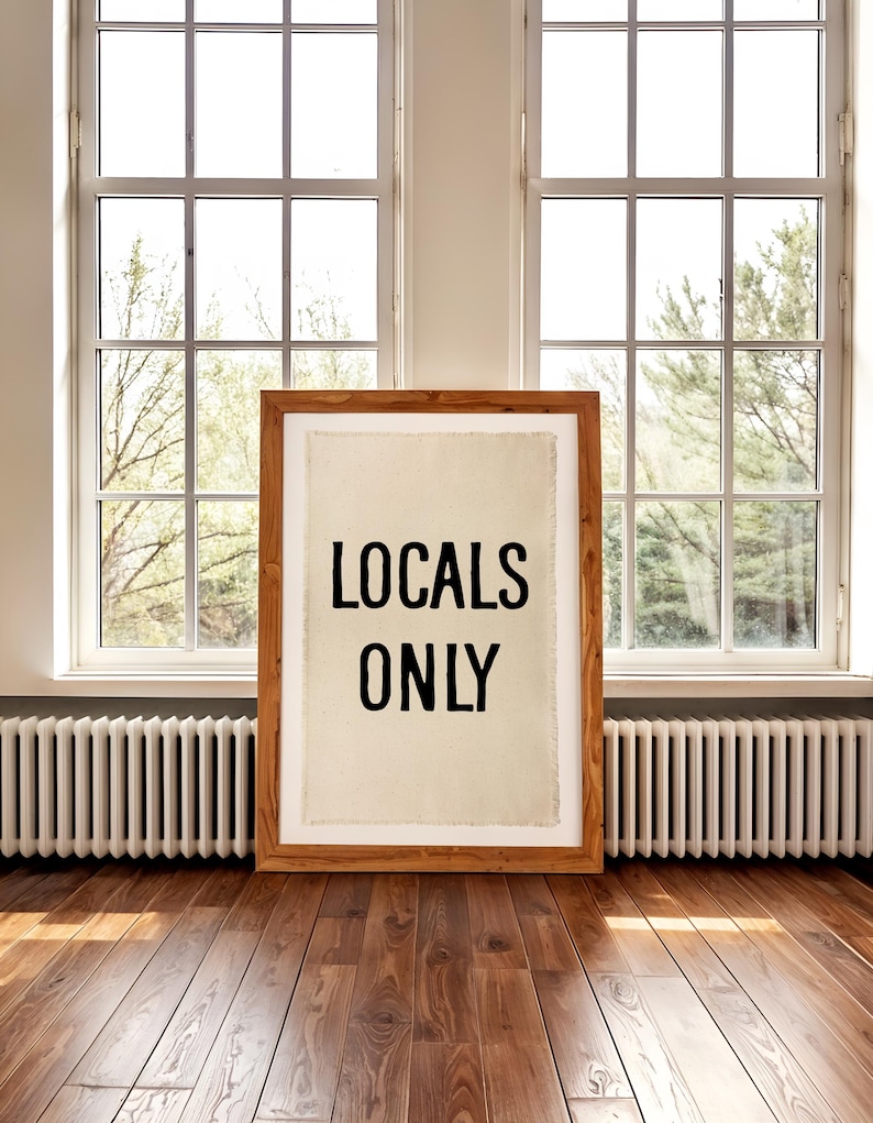 Pode incluir: Uma impress&atilde;o emoldurada em madeira com as palavras "LOCALS ONLY" em preto sobre um fundo creme. A impress&atilde;o est&aacute; colocada em um piso de madeira em frente a uma janela.