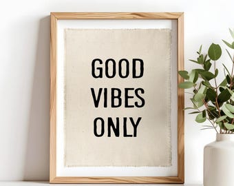 May include: A framed wall art piece featuring a light beige fabric insert with frayed edges. The text GOOD VIBES ONLY is printed in bold black capital letters in the center. The frame is made of light-colored wood with a visible grain pattern.