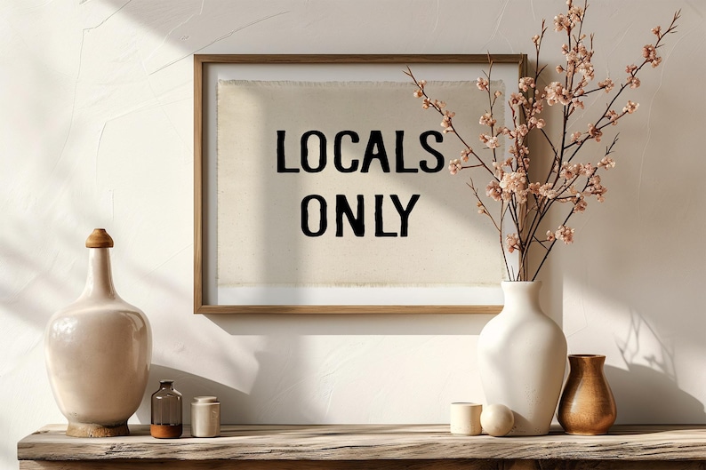 Pode incluir: Uma obra de arte emoldurada com o texto "LOCALS ONLY" em letras pretas. A obra de arte &eacute; exibida em uma superf&iacute;cie de madeira com vasos decorativos e um arranjo floral. A cena &eacute; banhada pela luz natural, criando um ambiente acolhedor e convidativo.