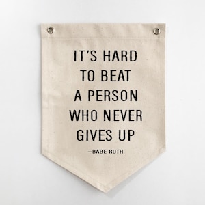 Op de afbeelding: Een rechthoekige wandbanner van gebroken wit canvas met een puntige onderkant en twee zilveren metalen ringen aan de bovenkant. De banner is voorzien van zwarte tekst die luidt: IT'S HARD TO BEAT A PERSON WHO NEVER GIVES UP -BABE RUTH.