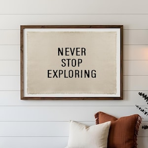 May include: A rectangular wall art piece featuring a beige fabric canvas with frayed edges, encased in a dark brown wooden frame. The canvas displays the black, sans-serif text "NEVER STOP EXPLORING" centered in three lines, creating a clear visual statement.