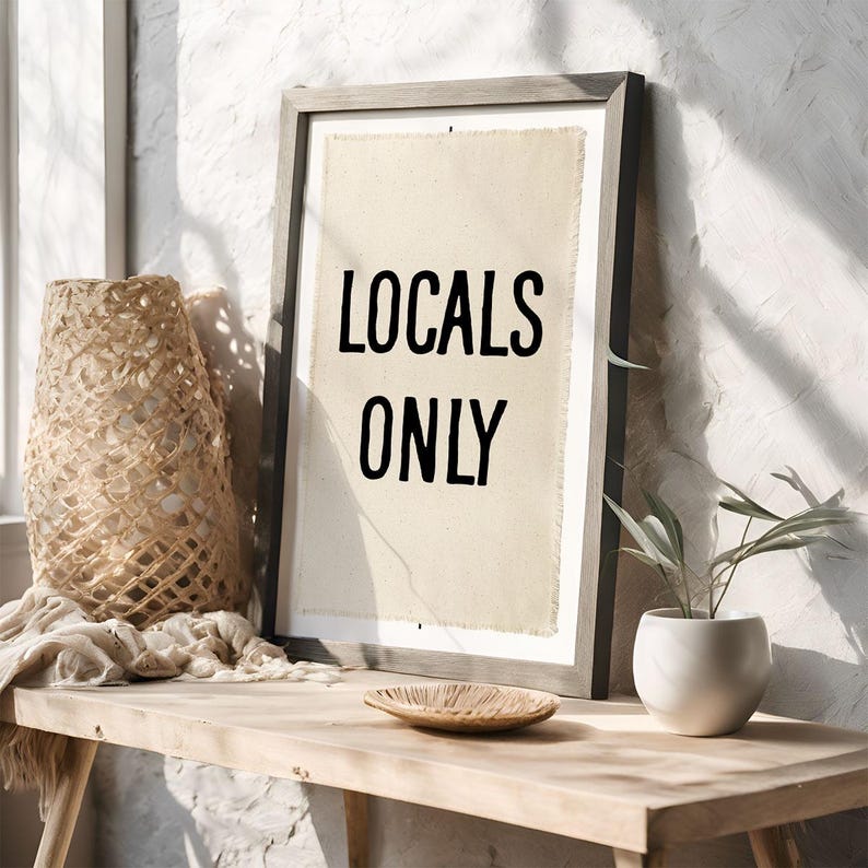 Pode incluir: Impress&atilde;o emoldurada com as palavras pretas "LOCALS ONLY" sobre um fundo bege. A moldura cinza est&aacute; sobre uma mesa de madeira, junto a uma cesta tecida, uma tigela pequena e uma planta em vaso, criando um ambiente r&uacute;stico.