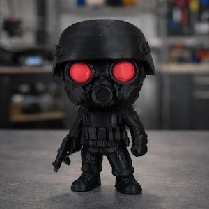 May include: A black figurine of a soldier wearing a helmet, gas mask, and tactical gear. The figure has glowing red eyes and is holding a rifle. The figurine is standing on a gray surface.