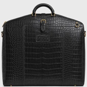 Puede incluir: Una funda para ropa negra con un patrón texturizado de piel de cocodrilo. El bolso incluye un asa superior, una correa vertical central con una hebilla dorada, herrajes de metal dorado y anillas en D laterales para enganchar una correa de hombro.
