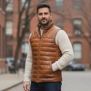 Könnte beinhalten: Eine braune Lederweste mit Steppdesign und Reißverschluss. Die Weste hat einen Stehkragen und zwei Vordertaschen. Die Person trägt einen cremefarbenen Pullover darunter. Der Hintergrund zeigt eine Stadtstraße mit Backsteingebäuden.