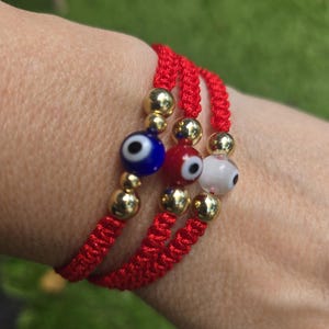 Puede incluir: Tres pulseras rojas tejidas con cuentas doradas y amuletos de ojo turco de cristal en azul, rojo y blanco. Las pulseras están apiladas una encima de otra.
