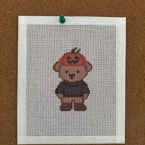 Könnte beinhalten: Eine Stickleinwand mit einem Kreuzstichmotiv eines hellbraunen Bären, der einen dunkelgrauen Pullover und einen leuchtend orangefarbenen Kürbishut mit einem geschnitzten Kürbisgesicht trägt. Das Design ist auf einem weißen Gitterstoff zentriert.