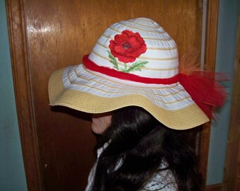 May include: A white and tan sun hat with a red poppy flower embroidered on the crown. The hat has a red ribbon around the brim and a red tulle bow on the side.