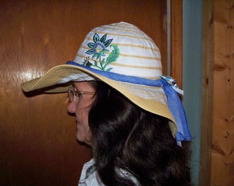 May include: A white and tan striped sun hat with a blue ribbon and a floral embroidery design. The hat has a wide brim and is made of straw.