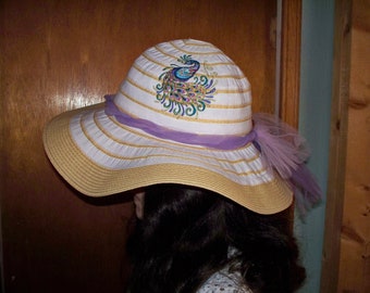 May include: A white and tan straw sun hat with a purple ribbon and a peacock design painted on the crown.