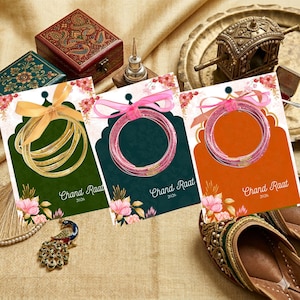 May include: Three cards displaying bangles in various colours, each adorned with a ribbon bow. The cards are green, teal, and orange, with the text "Chand Raat 2026". Decorative boxes and a peacock ornament are also visible.