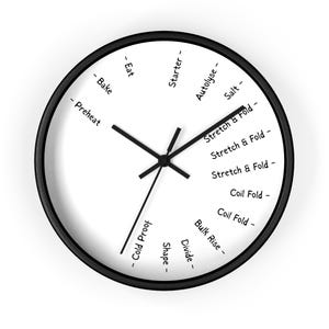 Puede incluir: Un reloj redondo con marco negro y esfera blanca. Las manecillas indican la hora. La esfera tiene texto negro que indica los pasos para hacer pan, como "Precalentar", "Stretch & Fold" y "Comer".
