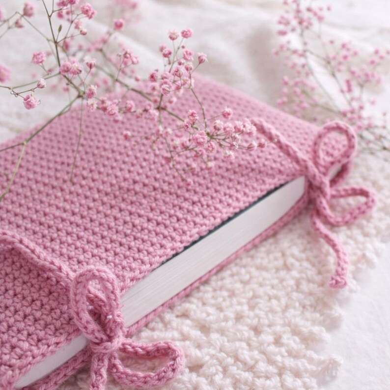 May include: A dusky pink crocheted book cover with a tie closure. The cover is wrapped around a book with white pages. Sprigs of pink flowers are arranged around the book, creating a soft, feminine aesthetic.