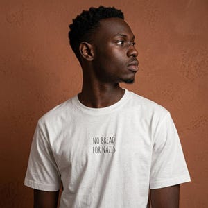 Könnte beinhalten: Weißes T-Shirt mit dem schwarzen Text "NO BREAD FOR NAZIS". Das T-Shirt wird von einer Person mit dunkler Haut und kurzen, dunklen Haaren getragen. Der Hintergrund ist eine strukturierte, braune Wand.