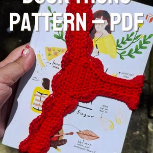 Könnte beinhalten: Ein rotes, gehäkeltes Buchzeichen ist auf einem Buch zu sehen. Das Buch hat Illustrationen und den Text "BOOK THONG PATTERN - PDF" in Weiß. Der Text "Instant Download" ist ebenfalls sichtbar.