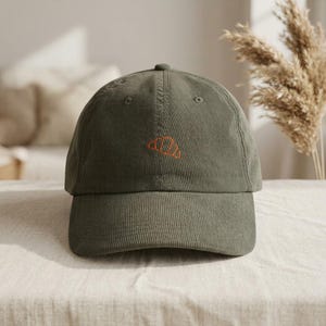 Op de afbeelding: Olijfgroene corduroy baseballcap met een gebogen klep. De pet heeft een geborduurd oranje croissantontwerp op de voorkant. De hoed staat op een neutraal gekleurd oppervlak.