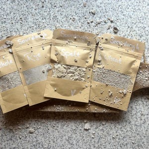 Könnte beinhalten: Vier braune Papiertüten mit Sichtfenster, die verschiedene Sauerteig-Starter enthalten. Jede Tüte ist mit weißer Schrift beschriftet, die Sourdough und dann Whole, Rye, Spelt oder Wheat lautet. Die Tüten liegen auf einem Holzbrett.