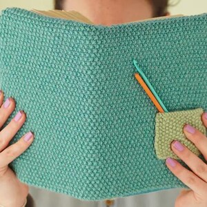 May include: A teal knitted book cover with a small pocket and two crochet hooks. The cover is made of a textured, turquoise yarn. The pocket is a lighter green colour. One crochet hook is orange, and the other is teal.