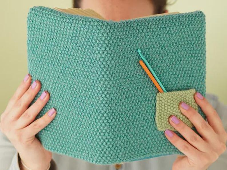 May include: A teal knitted book cover with a small pocket and two crochet hooks. The cover is made of a textured, turquoise yarn. The pocket is a lighter green colour. One crochet hook is orange, and the other is teal.