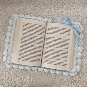 May include: An open book sits on a handmade crochet book cover. The cover is white with a scalloped edge and light blue detailing. A light blue ribbon is tied at the top. The book pages are visible.