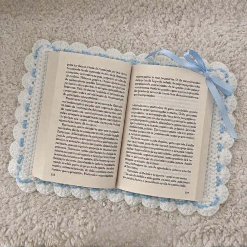 May include: An open book sits on a handmade crochet book cover. The cover is white with a scalloped edge and light blue detailing. A light blue ribbon is tied at the top. The book pages are visible.
