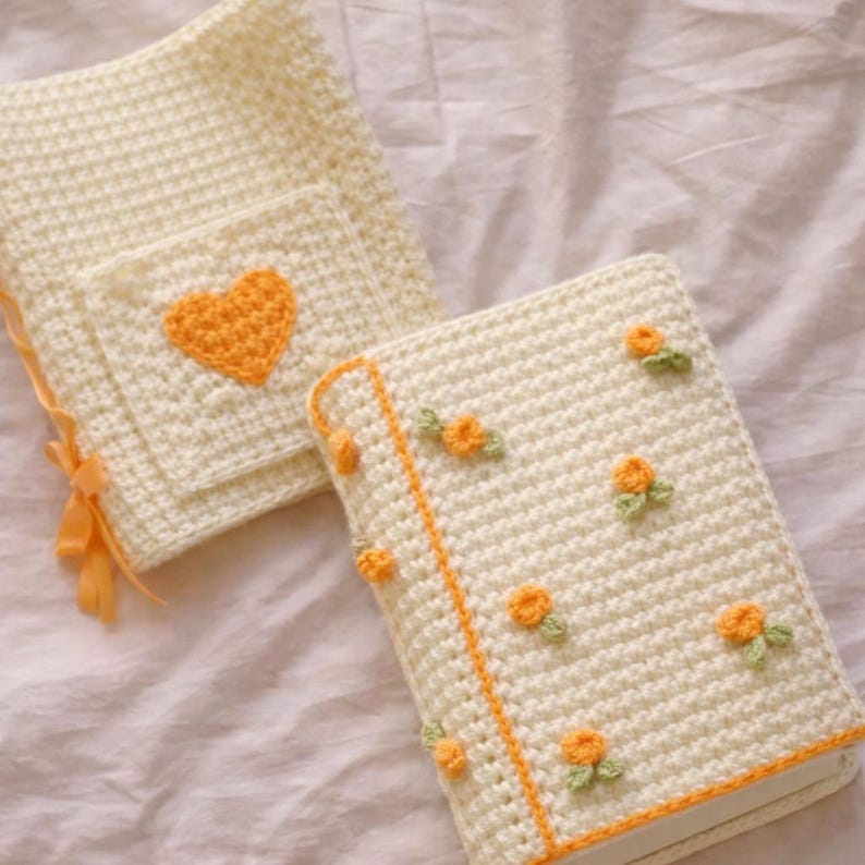 May include: Two cream-coloured crochet book covers. One has an orange heart on a pocket. The other has orange flowers and green leaves. Both have an orange border and accents. The covers are handmade and have a textured appearance.