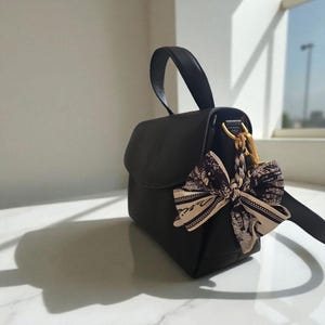May include: Black leather handbag with a top handle and a decorative bow. The bow is cream-coloured with a black pattern. The bag has a gold-coloured clasp and a detachable shoulder strap.