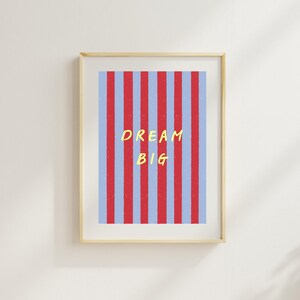 Könnte beinhalten: Ein gerahmter Wandkunstdruck mit vertikalen Streifen in abwechselndem Rot und Hellblau. Der Text DREAM BIG ist in gelben Großbuchstaben in der Mitte des Streifenmusters aufgedruckt und von einem hellen Holzrahmen umgeben.