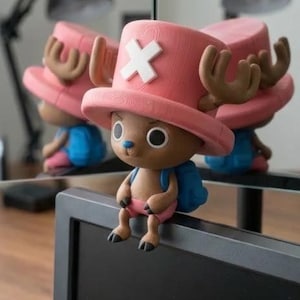 Peut inclure: Figurine d'un personnage de dessin animé avec un chapeau rose orné d'une croix blanche, des bois bruns et un sac à dos bleu. Le personnage est assis sur une surface noire. La figurine se reflète dans un miroir.