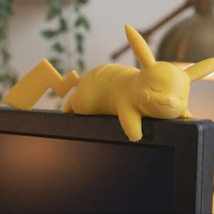Puede incluir: Una figura amarilla de Pikachu, con los ojos cerrados y una pose relajada, descansa sobre una superficie negra. La cola de la figura tiene forma de rayo. El fondo está borroso, con una lámpara y vegetación visible.