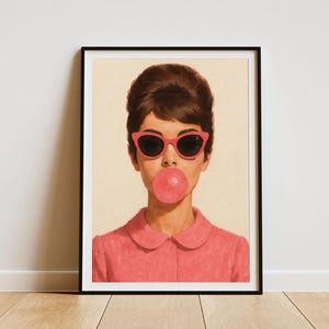 May include: Framed art print featuring a woman with dark hair wearing pink sunglasses and a pink collared shirt, blowing a bubblegum bubble. The artwork is displayed against a neutral background.