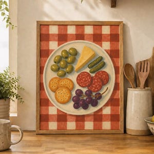 May include: A framed print featuring a plate of olives, cheese, crackers, salami, grapes, and pickles. The plate is set against a red and white checkered background. The artwork is displayed on a wooden countertop in a kitchen setting.