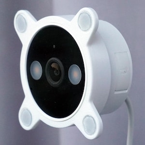 May include: A white security camera with a black lens and two orange lights. The camera has a circular design with four rounded corners and a white cable attached.