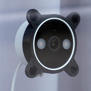 May include: A modern security camera with a black faceplate and white body. The camera has a circular design with a lens and two lights. The faceplate has four rounded corners. A white cable extends from the camera.