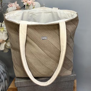 May include: A beige tote bag with white handles and a white interior. The bag features a textured, diagonal striped pattern in shades of brown and beige. A small silver tag with the letters "KPK" is sewn on the front.