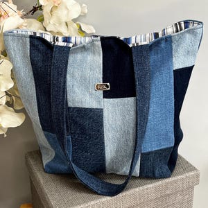 May include: A denim patchwork tote bag in shades of blue. The bag features a variety of denim squares, long straps, and a striped lining. A small gold-coloured metal tag with the letters "KPK" is attached to the front.