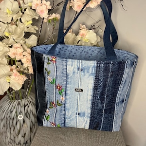 May include: A denim tote bag with dark blue handles and a floral embroidered design. The bag is made from repurposed denim and features a small metal tag with the initials "KPK". The interior lining has a blue floral pattern.