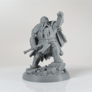 Op de afbeelding: Grijze miniatuurfiguur van een krijger met een bijl, met een opgeheven vuist. De figuur draagt een harnas en een cape, staand op een rotsachtige basis. Waarschijnlijk voor tabletop gaming of display, de figuur staat in een dynamische pose.